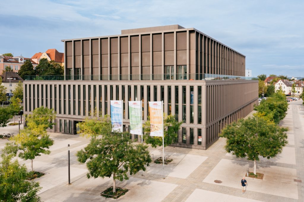 Stadthalle Reutlingen von außen mit grünen Bäumen im Vordergrund und drei farbigen Fahnen.