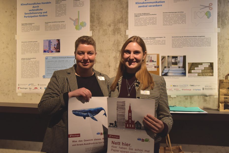 Im Vordergrund halten zwei Frauen, links Louisa Bahl und rechts Melanie Sinn, zwei Papiersparplakate in der Hand. Im Hintergrund sieht man Ausschnitte von Din-A1-Plakaten auf einer Betonwand.