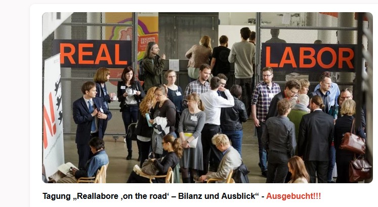 20-30 Menschen stehen oder sitzen in Kleingruppen zusammen und unterhalten sich. Im Hintergrund ist der Schriftzug "Reallabor" in orange auf braun-schwarzem Hintergrund zu sehen.