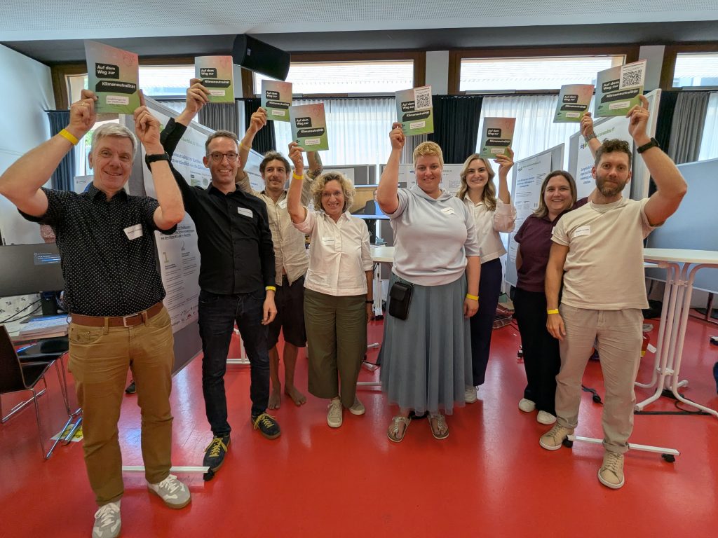 Gruppenbild des Teams des Klima-RT-LABs mit insgesamt 8 Personen, die alle die bunte Abschlussbroschüre des Reallabors über den Kopf strecken und grinsen und vor ihrem Stand mit Stellwänden stehen.