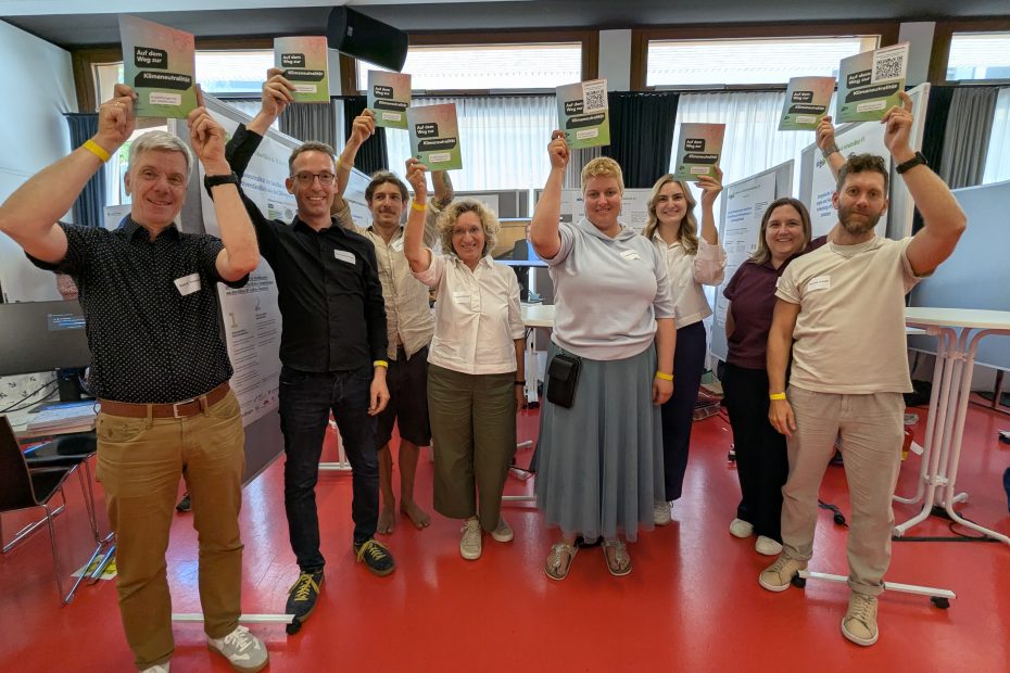 Gruppenbild des Teams des Klima-RT-LABs mit insgesamt 8 Personen, die alle die bunte Abschlussbroschüre des Reallabors über den Kopf strecken und grinsen und vor ihrem Stand mit Stellwänden stehen.
