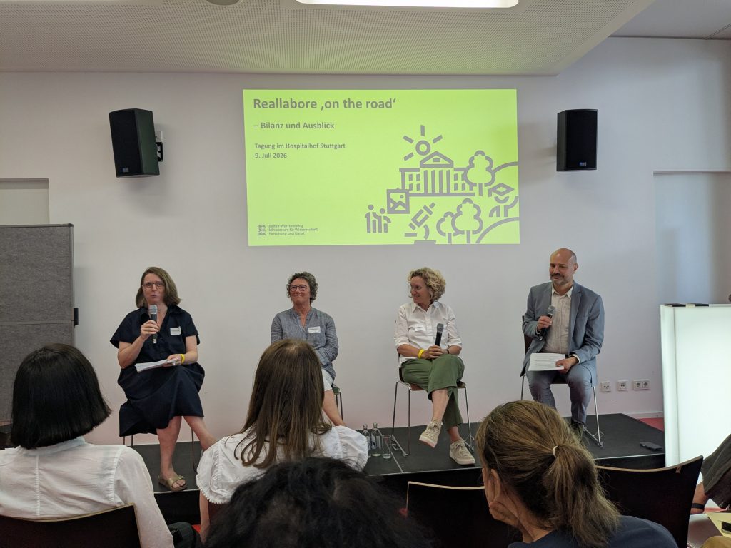 Podiumsdiskussion. Im Vordergrund einige Köpfe von hinten, auf der Bühne sitzen drei Frauen und ein Mann, dahinter eine gelbe PowerPoint-Folie.